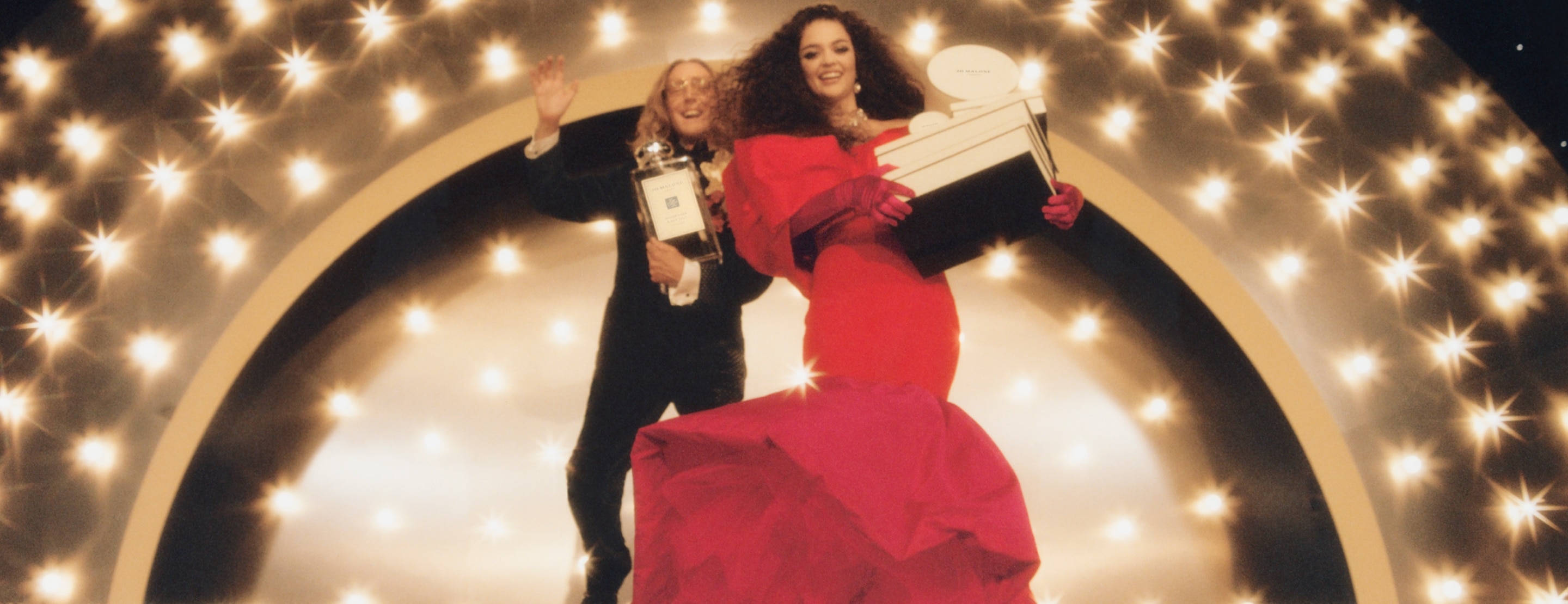 Two elegantly dressed individuals, holding an oversized cologne and box against a dazzling backdrop. 