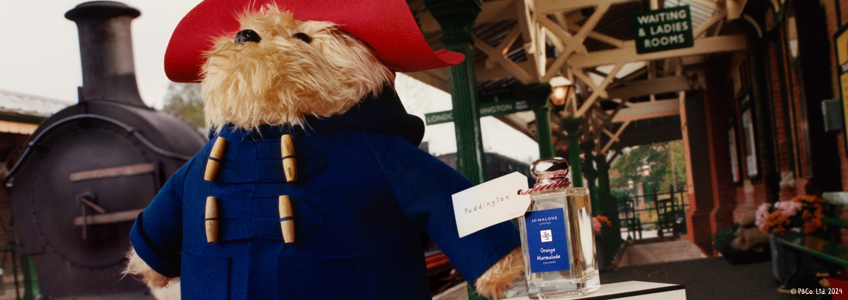 Paddington Bear and Orange Marmalade Cologne with a Paddington tag hanging from the clear, glass bottle in a train station 